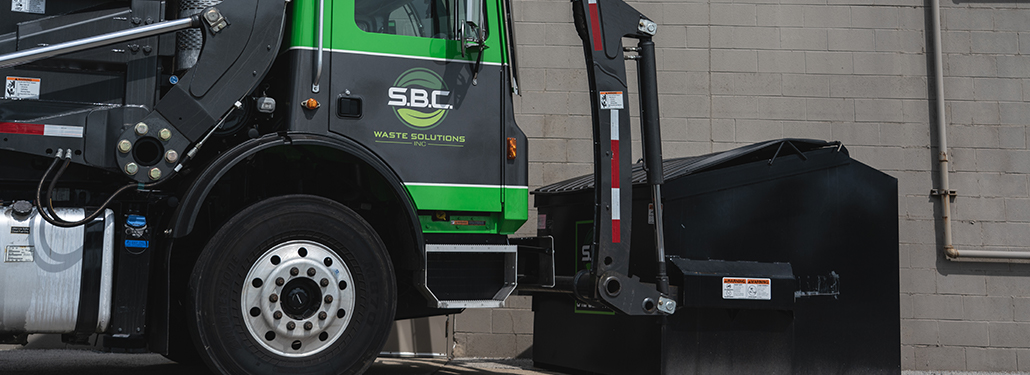 A dump truck beginning the process of lifting a front load dumpster, with the dumpster still on the ground.